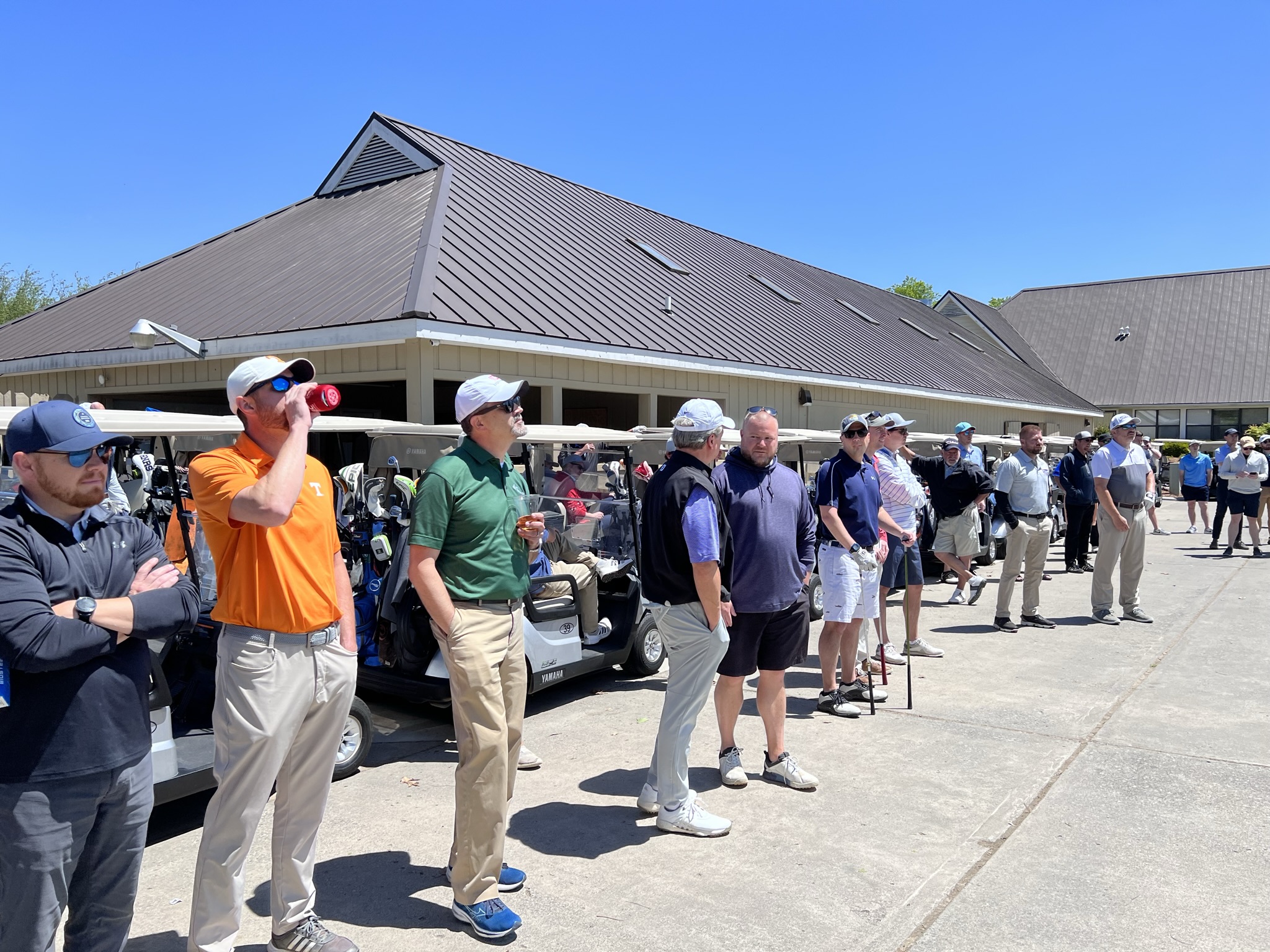 Group of men standing near golf carts outside a clubhouse on a sunny day.